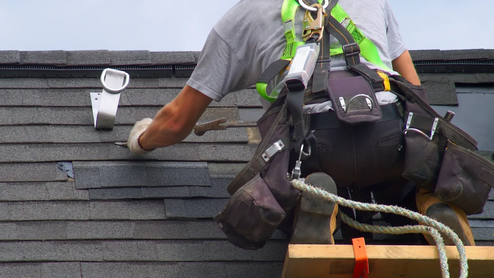 Roofer wearing a safety harness fitting a shingle on a gray roof
